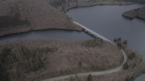 Drone-shot-in-Luxemburg-near-the-river-Lac-de-la-Haute-Sure-at-a-nature-park-Öewersauer-on-a-cloudy-day-with-cars-driving-on-the-road-near-the-forest-and-trees-and-a-bridge-in-the-background-LOG