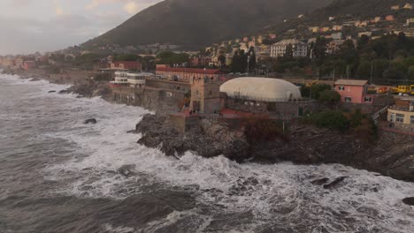 The-rocky-coastline-and-buildings-of-genoa,-italy,-with-waves-crashing-against-the-shore-at-dusk,-aerial-view