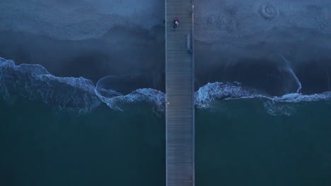 Top-down-drone-footage-of-a-pier-with-a-fisherman-walking-and-waves-crashing