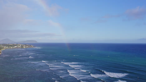 Drohnenaufnahmen-Fangen-Einen-Regenbogen-Im-Meeresnebel-Entlang-Der-Küste-Von-Oahu-Auf-Den-Hawaii-Inseln-Ein,-Mit-Vulkanbergen-Am-Horizont-Und-Weißen-Wellen,-Die-An-Die-Küste-Rollen