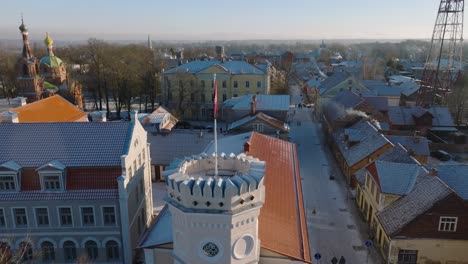 Aerial-establishing-view-of-Kuldiga-Old-Town-,-houses-with-red-roof-tiles,-tower-with-Kuldiga-flag,-sunny-winter-day,-travel-destination,-wide-drone-orbit-shot