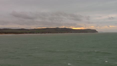 Coastal-view-of-the-French-shoreline-from-the-English-Channel-at-dusk,-with-gentle-waves-and-cloudy-skies