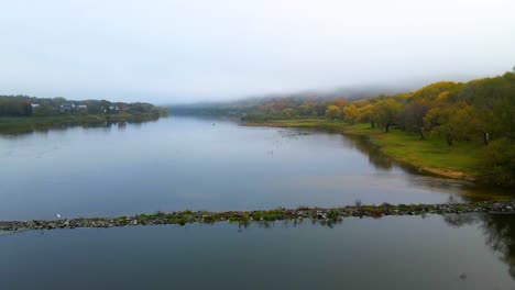 Drone-shot-of-stone-pier-on-the-Nemunas-river-with-dense-colorful-forest-and-houses-on-the-banks-at-sunrise-in-autumn