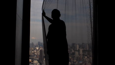 Silhouette-of-a-man-looking-out-the-window,-drawing-the-curtains-closed,-obscuring-the-panoramic-view-of-the-city-metropolis