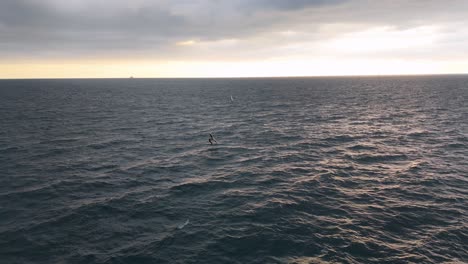 Solo-wind-surfer-in-vast-ocean-with-distant-ship,-cloudy-skies-over-Genoa,-Italy,-sunset-on-horizon