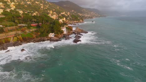 Nervi-in-genoa,-italy-with-waves-crashing-on-rocky-shore,-houses-nestled-on-hillside,-aerial-view