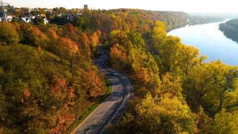 Droneshot-of-a-car-driving-on-winding-road-adjacent-to-railway-alongside-Nemunas-River-in-Kaunas,-Lithuania,-zoom-in-shot