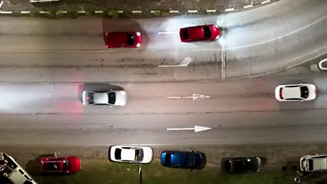 Car-headlights-shine-as-they-cross-over-faded-white-arrow-in-asphalt,-drone-at-night
