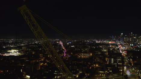 Aerial-tracking-shot-of-a-construction-crane-with-night-lit-LA-cityscape-background