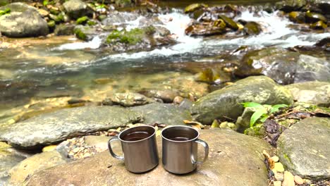 B-roll-footage-of-two-coffee-mugs-full-of-fresh-coffee-placed-next-to-a-running-riverbed-in-nature