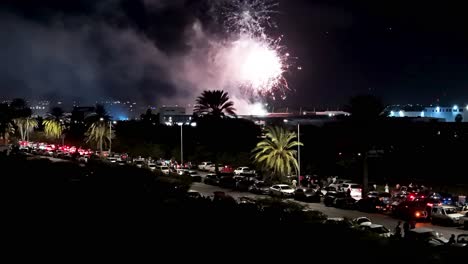 Aerial-fireworks-launch-high-into-sky-above-palm-trees-tropical-promenade-as-cars-line-road-to-watch-plume-of-smoke-and-sparkling-light