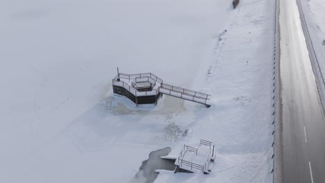 Drone-captures-a-snowy-road-with-a-frozen-lake-and-dam-structure-in-a-serene-Baltic-setting