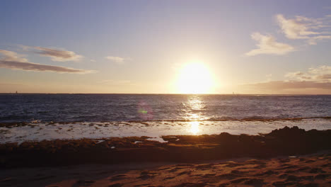 a-brilliant-white-light-sun-sets-behind-the-Pacific-Ocean-from-a-sandy-beach-on-Diamond-Head-Hawaii-Oahu-as-the-Pacific-waves-roll-onto-the-sandy-beach