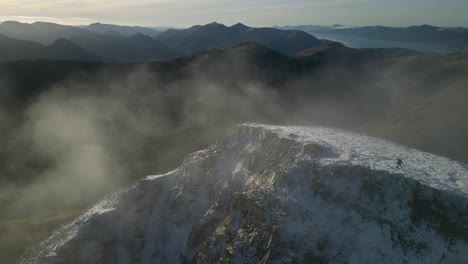 Caminante-Alcanzando-La-Cumbre-De-Una-Montaña-Nevada-Con-Nubes-Arremolinándose-Stob-Ban-Highlands-Escocia