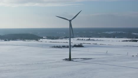 Wind-turbine-slowly-rotates-in-a-snowy-landscape-in-slow-motion,-with-a-backdrop-of-distant-trees-and-fields