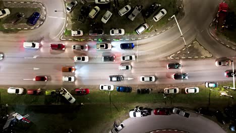 Cars-lineup-at-traffic-light-crowded-with-vehicles-parked-on-grass-median,-aerial-top-down-at-night