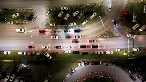 Cars-stop-at-traffic-light-rounding-bend-at-night-with-vehicles-parked-on-grass,-aerial