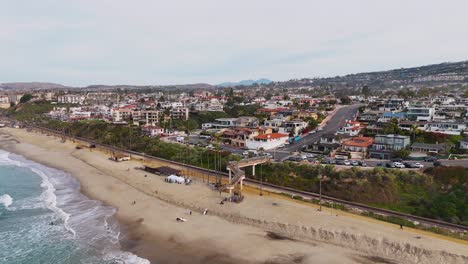 Aerial-View-of-California-beach-town-with-surfers-crossing-bridge-over-train-tracks