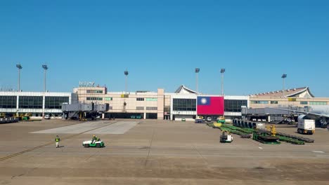 Outside-view-of-Taiwan-Taoyuan-International-Airport-terminal-building-with-Taiwanese-flag,-workers,and-transportation-vehicles-on-the-runway