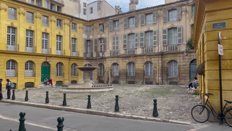Gelbe-Gebäude-Säumen-Einen-Kopfsteinpflasterplatz-In-Aix-en-Provence,-Tagsüber-Schlendern-Menschen-Und-Sitzen-In-Der-Nähe-Eines-Klassischen-Brunnens