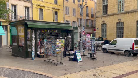 Newsstand-filled-with-magazines-and-postcards-in-Aix-en-Provence,-with-people-and-vehicles-in-background