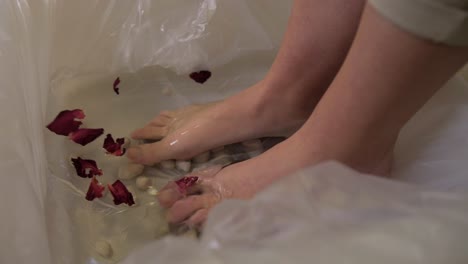 woman-stepping-barefoot-in-water-bucket-with-petals-slow-motion,-spa-pampering