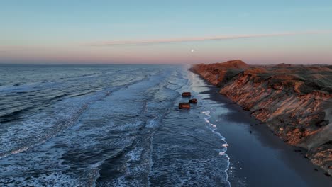 Aerial-view-Sea-waves-are-washing-on-the-beach,-high-dunes-have-formed,-the-remains-of-war-bunkers-are-on-the-beach,-and-the-sun-is-setting-on-a-winter-evening