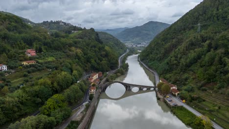A-serpentine-river-with-a-bridge-in-tuscany,-lush-greenery-on-both-sides,-cloudy-day,-aerial-hyperlapse