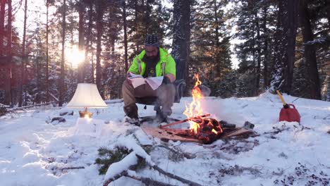 Man-sitting-on-a-chair-next-to-a-fire-and-lamp-in-a-forest-covered-with-snow