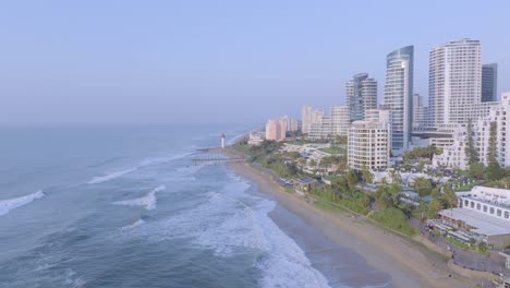 Umhlanga-coastline-with-waves-and-modern-buildings,-clear-sky,-aerial-view