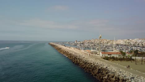 Puerto-Deportivo-De-Puerto-Banús-En-Marbella-Con-Embarcaciones-De-Recreo-Y-Una-Lancha-Rápida-En-Movimiento,-Cielo-Despejado,-Vista-Aérea