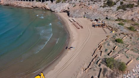 Yellow-excavator-working-on-idyllic-beach-near-port-ginesta-beach-in-barcelona,-spain-with-clear-waters-and-sandy-shore,-aerial-view