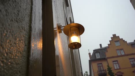 Warm-street-lamp-glowing-on-a-rainy-day,-droplets-on-wall,-overcast-sky,-European-architecture-in-the-background