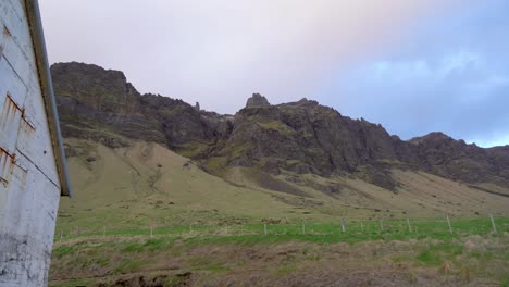 Rugged-Icelandic-landscape-with-green-meadows-and-imposing-mountain-peaks-under-a-cloudy-sky,-side-view-of-a-rustic-building