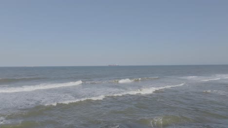 Gentle-waves-lapping-at-the-shore-with-a-distant-cargo-ship-on-the-horizon,-clear-blue-sky