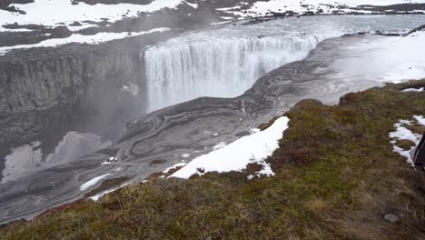 Impresionante-Vista-De-Una-Majestuosa-Cascada-En-Islandia-Con-Patrones-Arremolinados-En-El-Río,-Parches-De-Nieve-Y-Cielos-Nublados