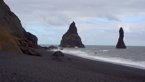 Black-sand-beach-with-towering-sea-stacks-under-a-cloudy-sky-in-Iceland,-dramatic-coastal-landscape
