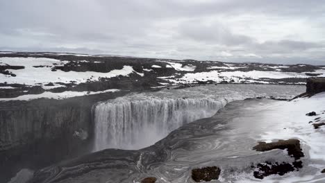 Majestätischer-Wasserfall-In-Island-Mit-Wirbelnden-Mustern-Im-Vordergrund,-Schneeflecken-Und-Bewölktem-Himmel