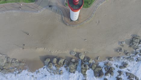 The-red-and-white-umhlanga-lighthouse-on-sandy-beach-with-waves-crashing-on-rocks,-aerial-view