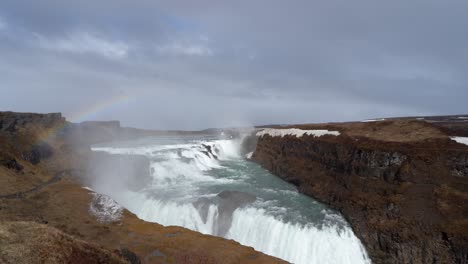 Majestic-Gullfoss-waterfall-with-vibrant-rainbow-in-Iceland,-cloudy-skies-overhead,-winter-season
