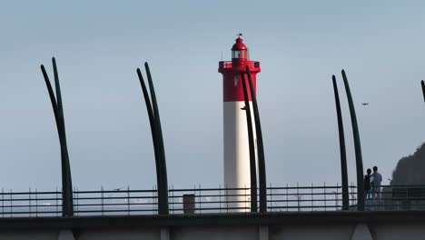Red-and-white-lighthouse-at-Umhlanga-with-silhouettes-of-a-couple-on-the-bridge,-clear-skies