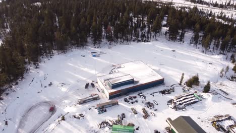 Aerial-view-of-a-construction-site-with-a-blue-warehouse-building-in-the-center-surrounded-by-snow-and-dense-forest