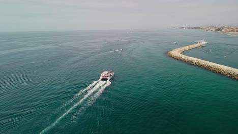 A-boat-cruising-near-the-breakwater-of-puerto-banus,-marbella-on-a-sunny-day,-aerial-view