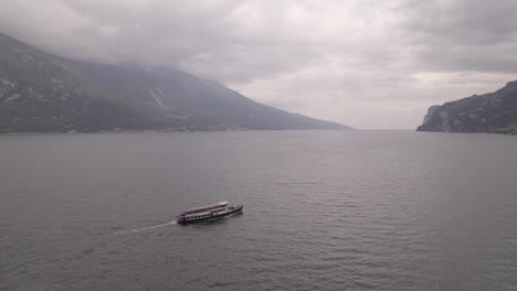 Droneshot-of-a-ferry-boat-going-to-the-city-Limone-Italy-on-Lake-Garda-on-a-grey-day-with-mountains-and-water-LOG