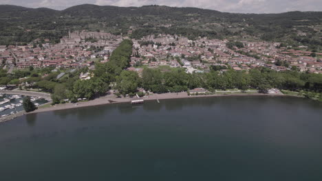 Drone-shot-flying-over-Lake-Bolsena-in-Italy-coming-from-the-water-into-the-old-medieval-city-with-a-castle-and-old-buildings-on-the-hill-on-a-sunny-day-LOG