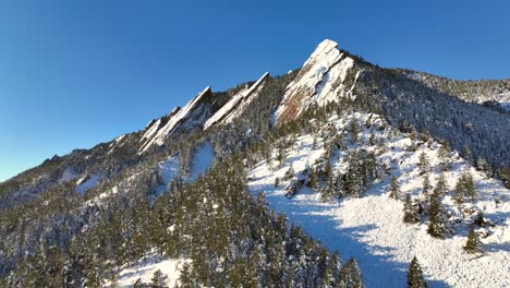 Slow-drone-reverse-above-the-tops-of-trees-on-a-bright-winter-day-with-snow-covering-the-flatiron-mountains-in-chautauqua-Park