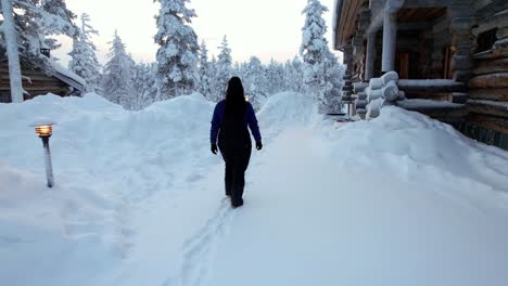 Chica-Caminando-Hacia-La-Cabaña-En-El-País-De-Las-Maravillas-Del-Invierno-Cubierto-De-Nieve-En-Laponia,-Finlandia,-El-Círculo-Polar-ártico