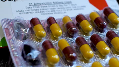 Eine-Nahaufnahme-Von-Amoxicillin-Kapseln,-Während-Die-Dunkelroten-Und-Gelben-Bestandteile-Sorgfältig-In-Ihrer-Originalverpackung-Angeordnet-Werden-Und-Zwei-Pillen-Herausgenommen-Werden