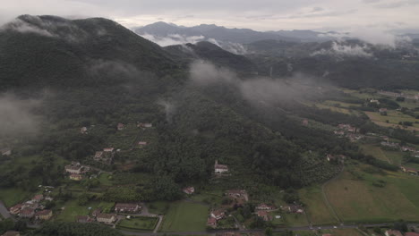 Drone-shot-of-clouds-moving-past-the-camera-in-the-hills-and-mountains-in-Italy-on-a-cloudy-grey-day-with-villages-and-houses-down-on-the-ground-in-between-green-fields-LOG