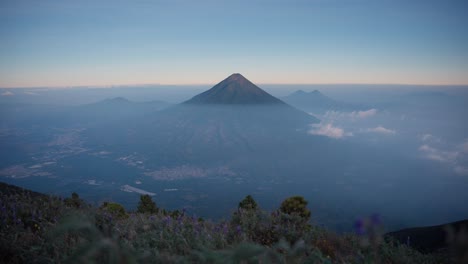 Der-Vulkan-Agua-Thront-über-Dem-Nebligen-Tal,-Blick-Auf-Den-Sonnenuntergang,-Unten-Antigua,-Violette-Blumen-Im-Vordergrund